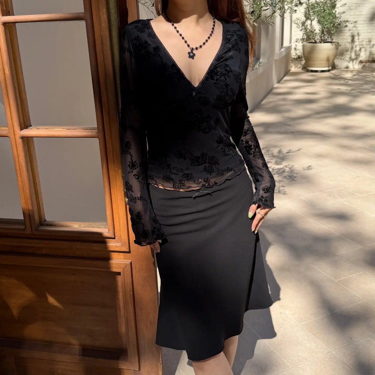 Model wearing floral flocked v neck bow mesh long sleeve top with black midi skirt, outdoor full view