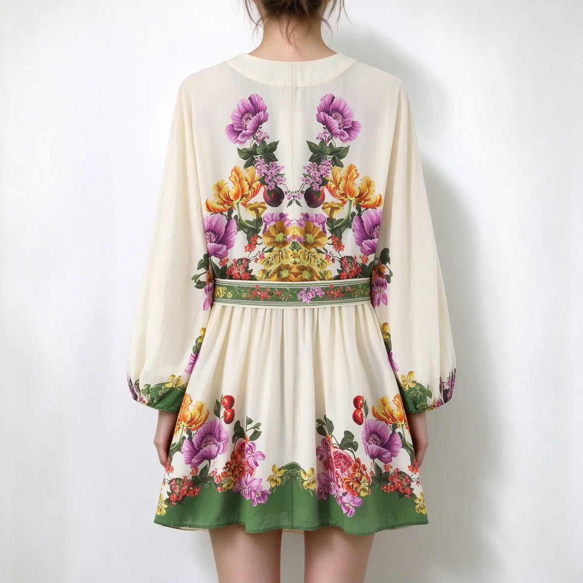 Model back view, boho mini dress with vibrant floral and fruit print, long balloon sleeves, matching printed waist tie, green hem trim, off-white base