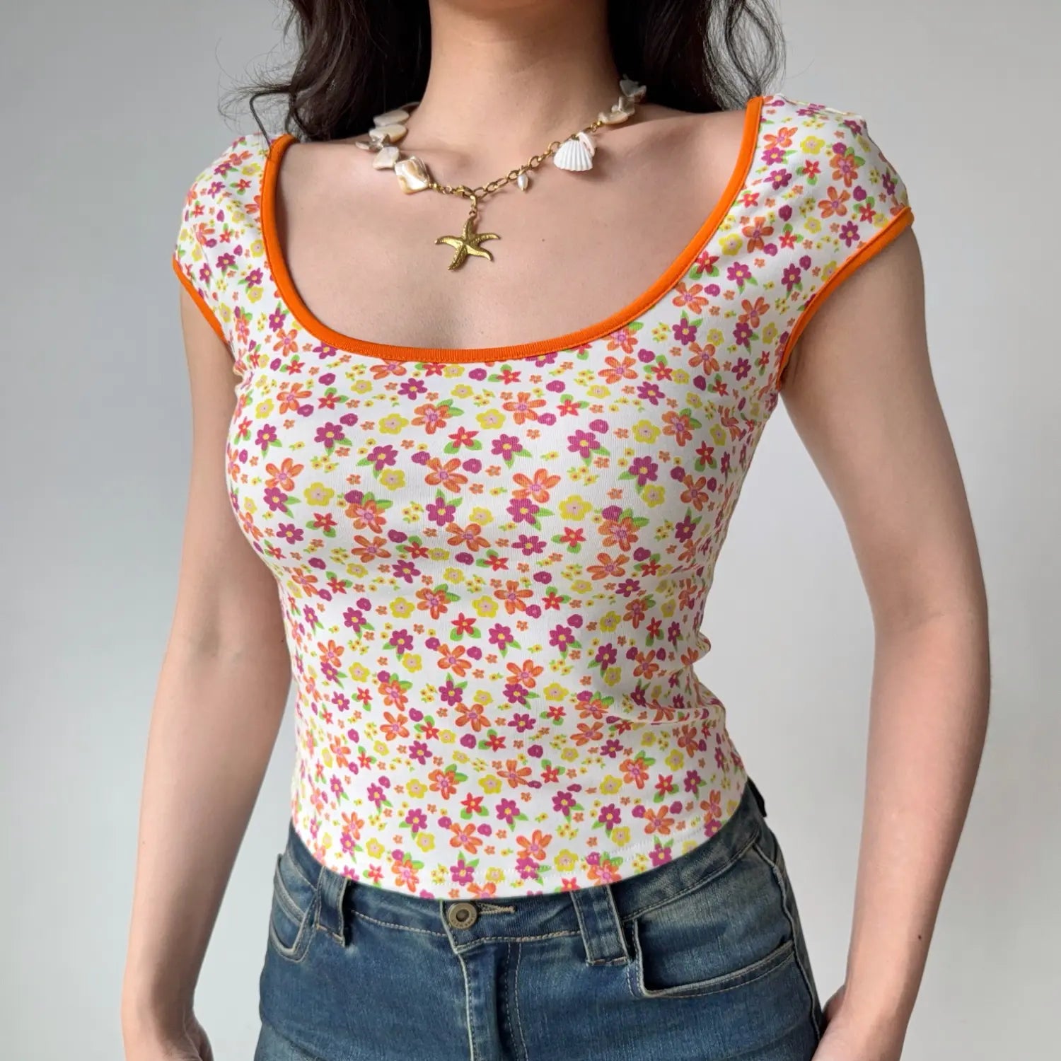 Lifestyle shot of a woman wearing a fitted floral print top with orange trim and a shell necklace