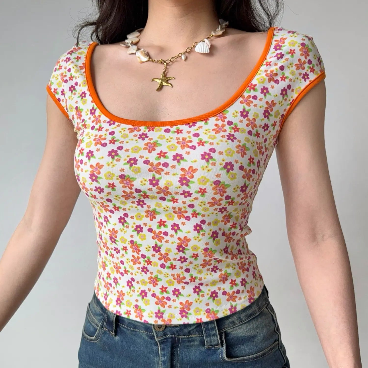 Lifestyle shot of a woman wearing a fitted floral print top with orange trim and a shell necklace, arms relaxed at sides