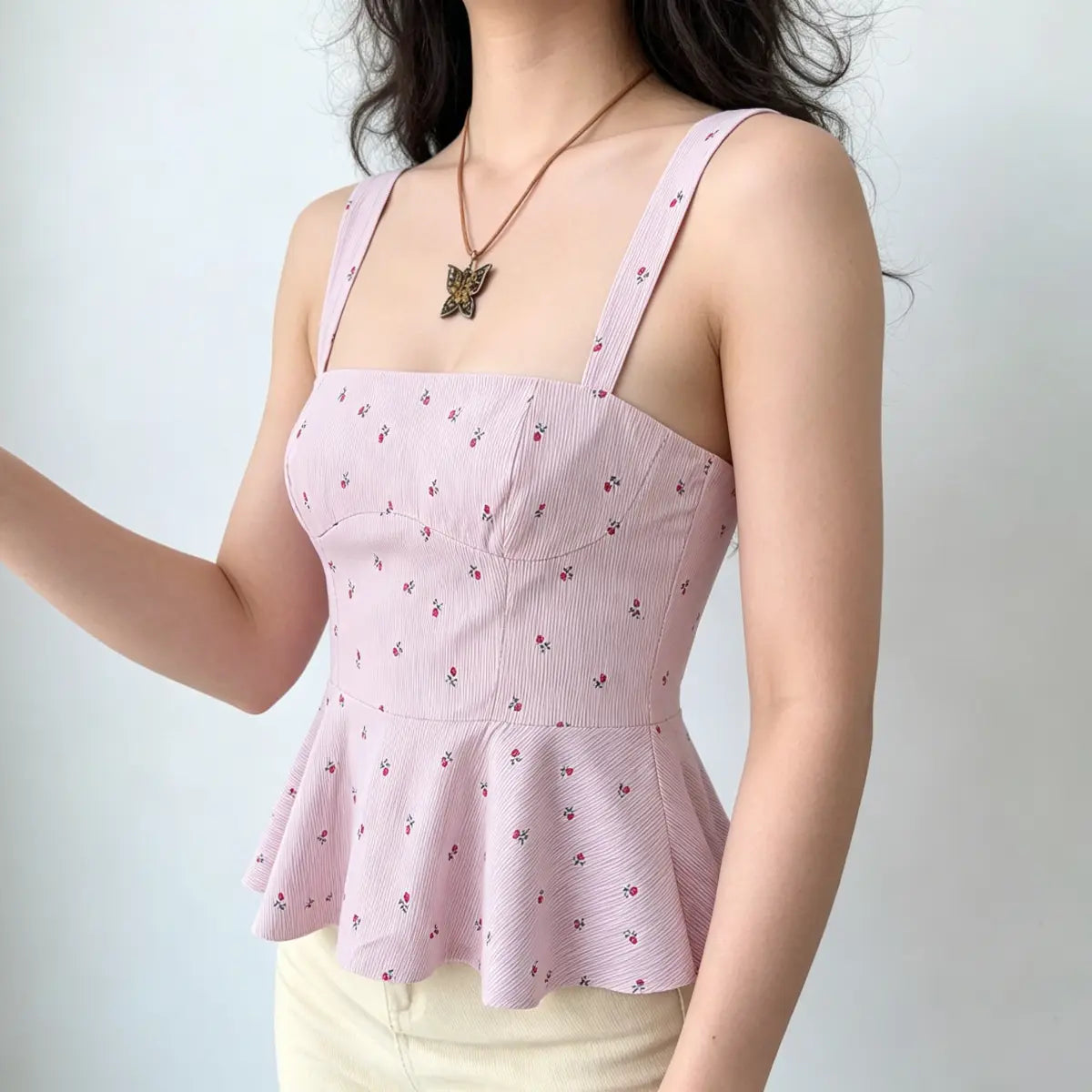 Model with arm raised, wearing a light pink floral peplum cami top, emphasizing the fitted bodice and flowy peplum hem.