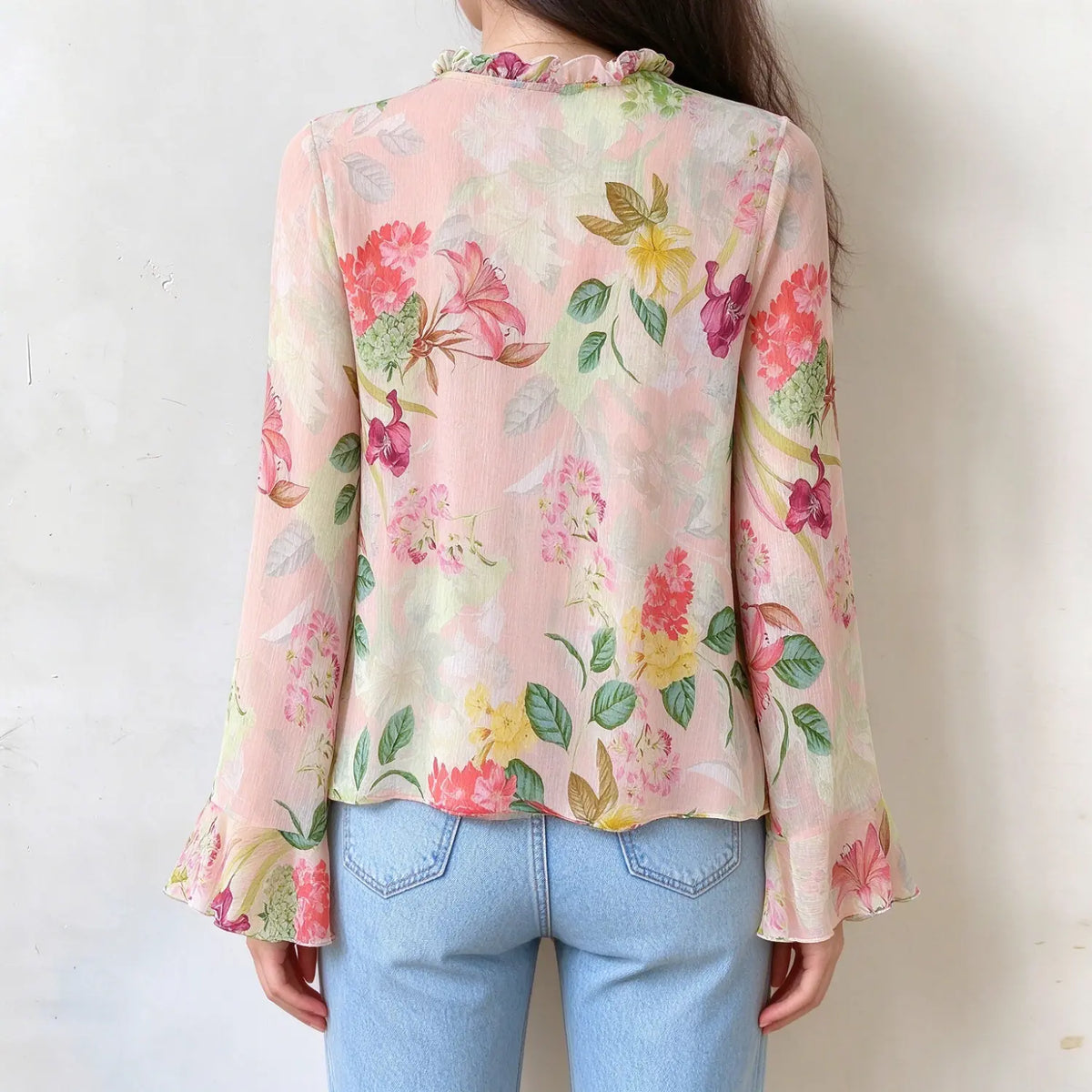 Female model back view of pink floral chiffon blouse, full back floral print, ruffle hem on flared long sleeves