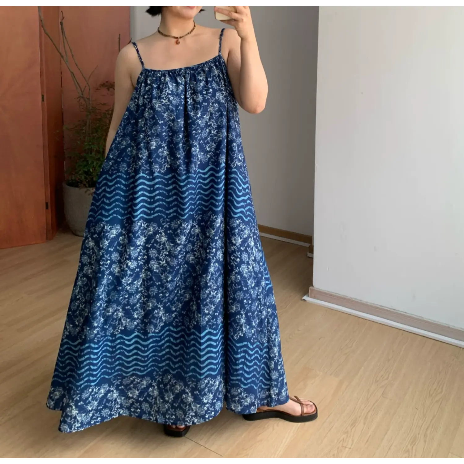 Full spread front indoor mirror shot of ultra-loose A-line batik slip maxi dress, deep indigo base with alternating white floral and pale blue horizontal wave stripe print panels, gathered square neck thin spaghetti straps, extra wide flowy floor-length skirt, brown thong slide sandals, simple potted plant room backdrop