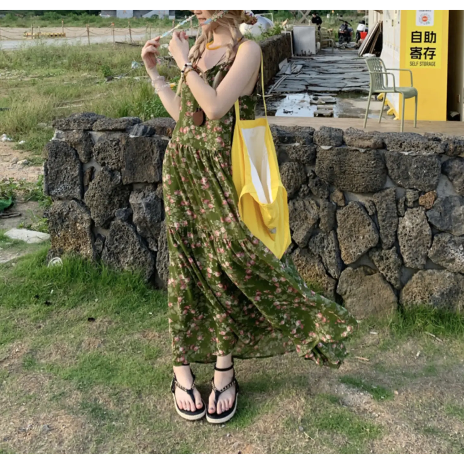 Windy outdoor stone wall lifestyle shot of dark green pink ditsy rose tier maxi slip dress, flowy ruffled skirt hem, black chain detail platform flip flops, large yellow canvas shoulder bag