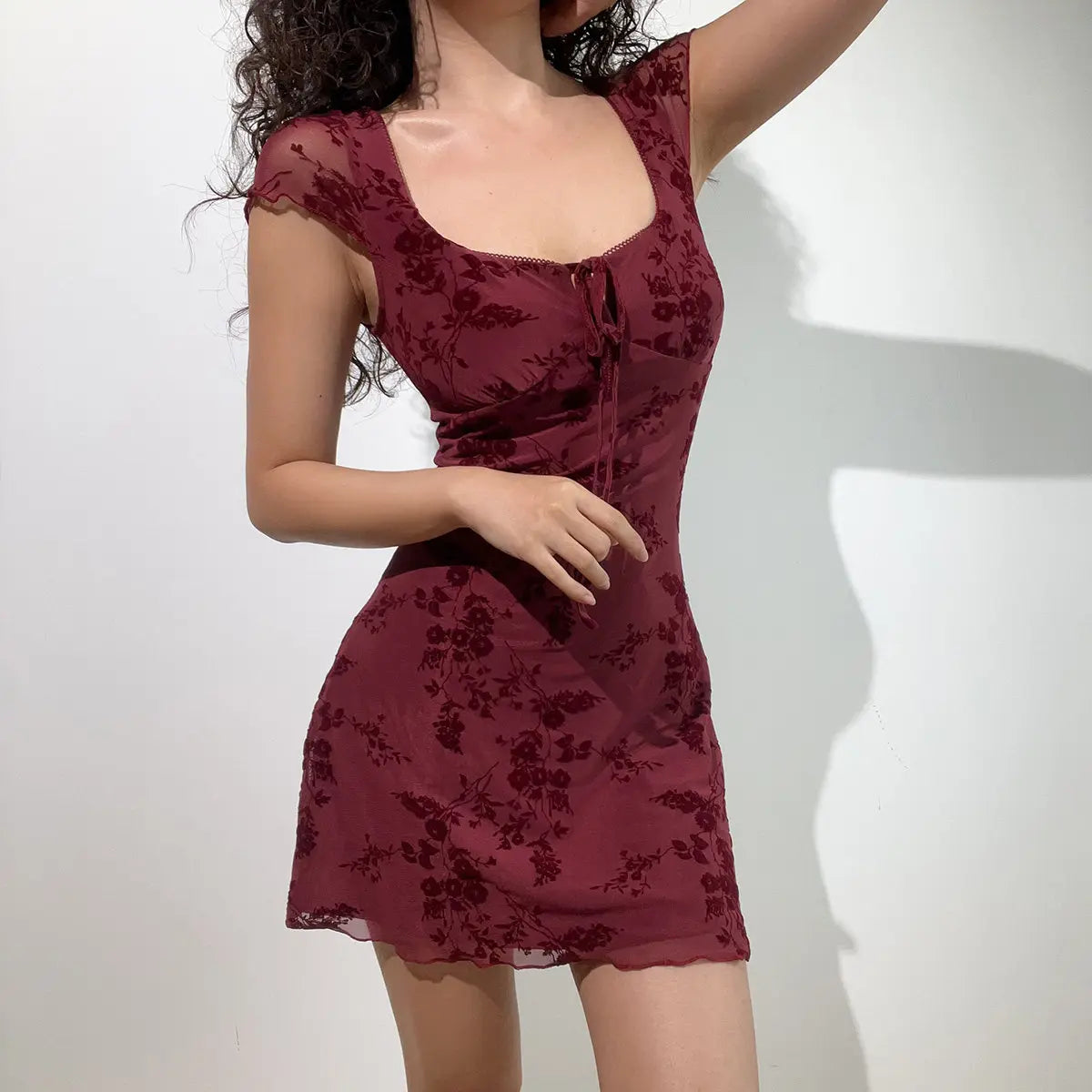 Full front view of a burgundy flocked floral mesh mini dress, featuring the square neckline, front tie, and flowy hem.
