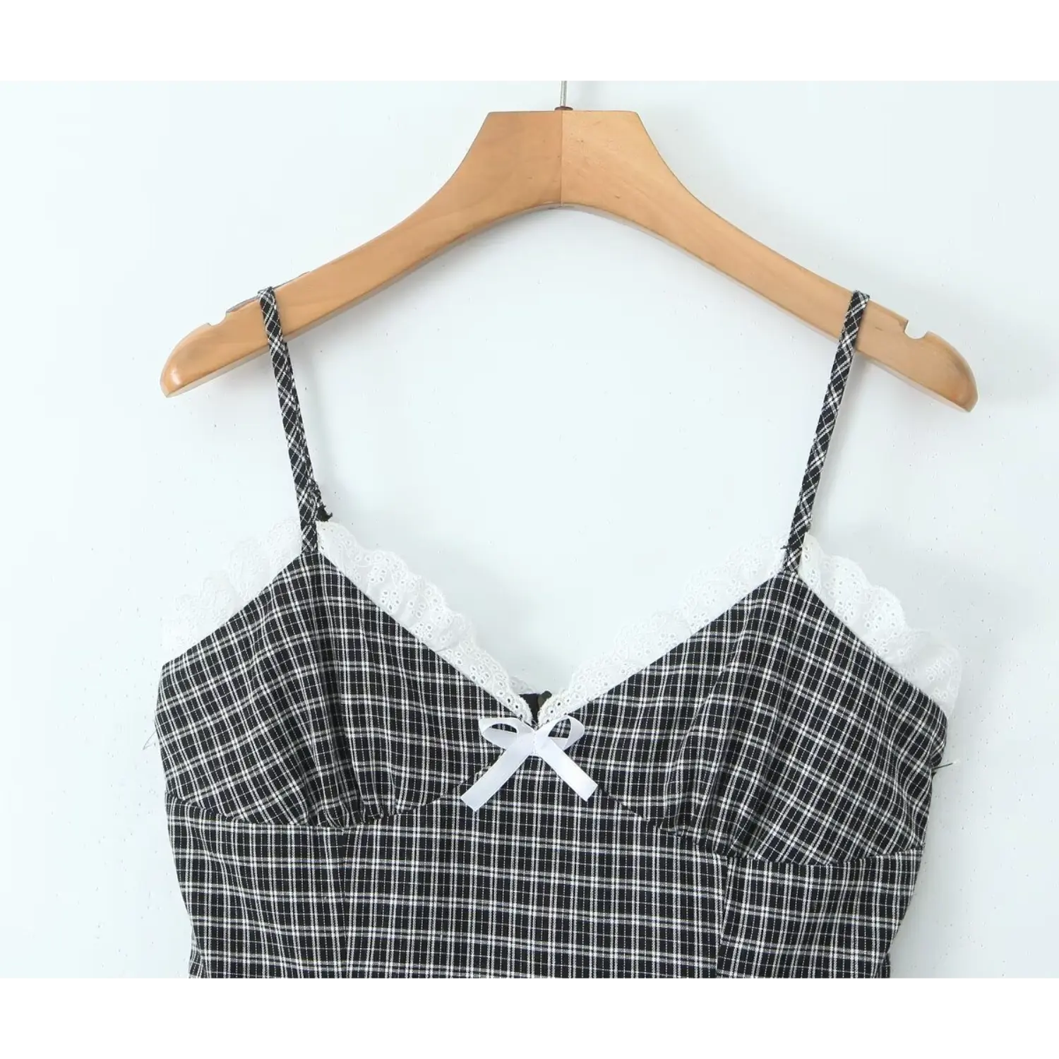 Close‑up bodice shot of black gingham cami dress hanging on wooden hanger, v‑neck white eyelet lace trim, small white satin bow