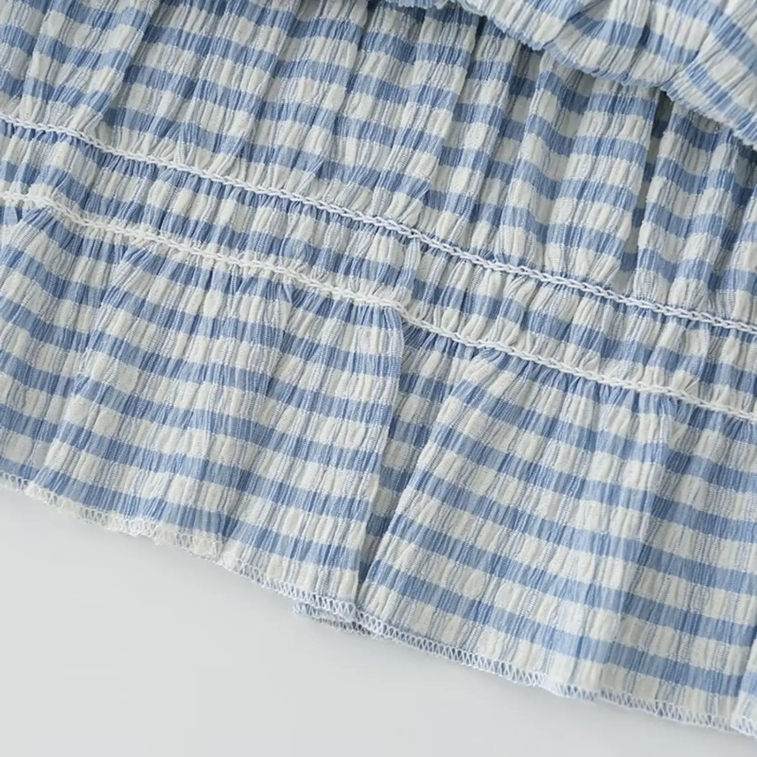 Macro stitching detail of peplum ruffle hem, uniform elastic smock seams, finished narrow hem edge, light blue gingham crinkle cloth