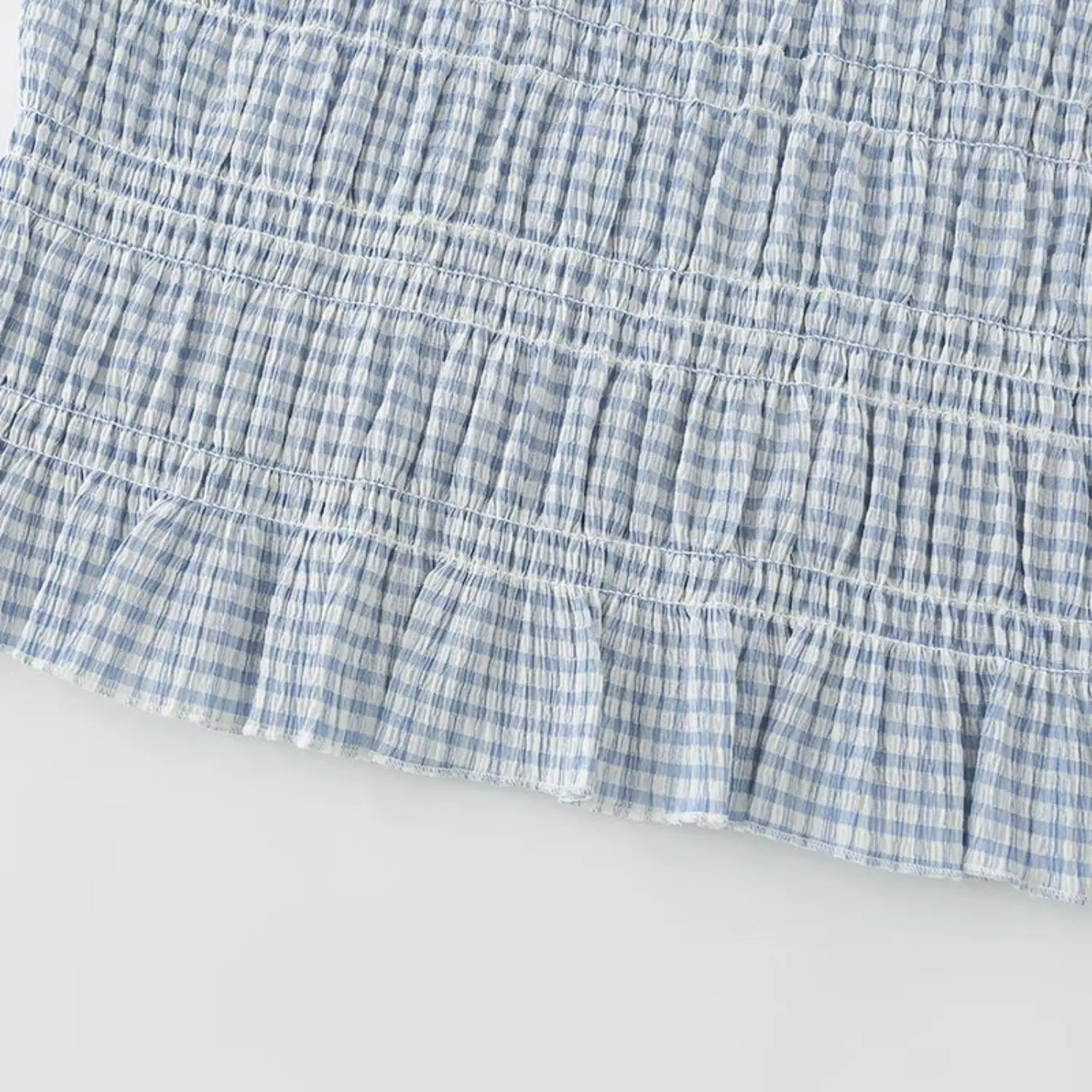 Closeup of light blue gingham peplum hem, multi-line horizontal elastic smocked stitching, ruffle flared hem, crinkle textured tiny check fabric