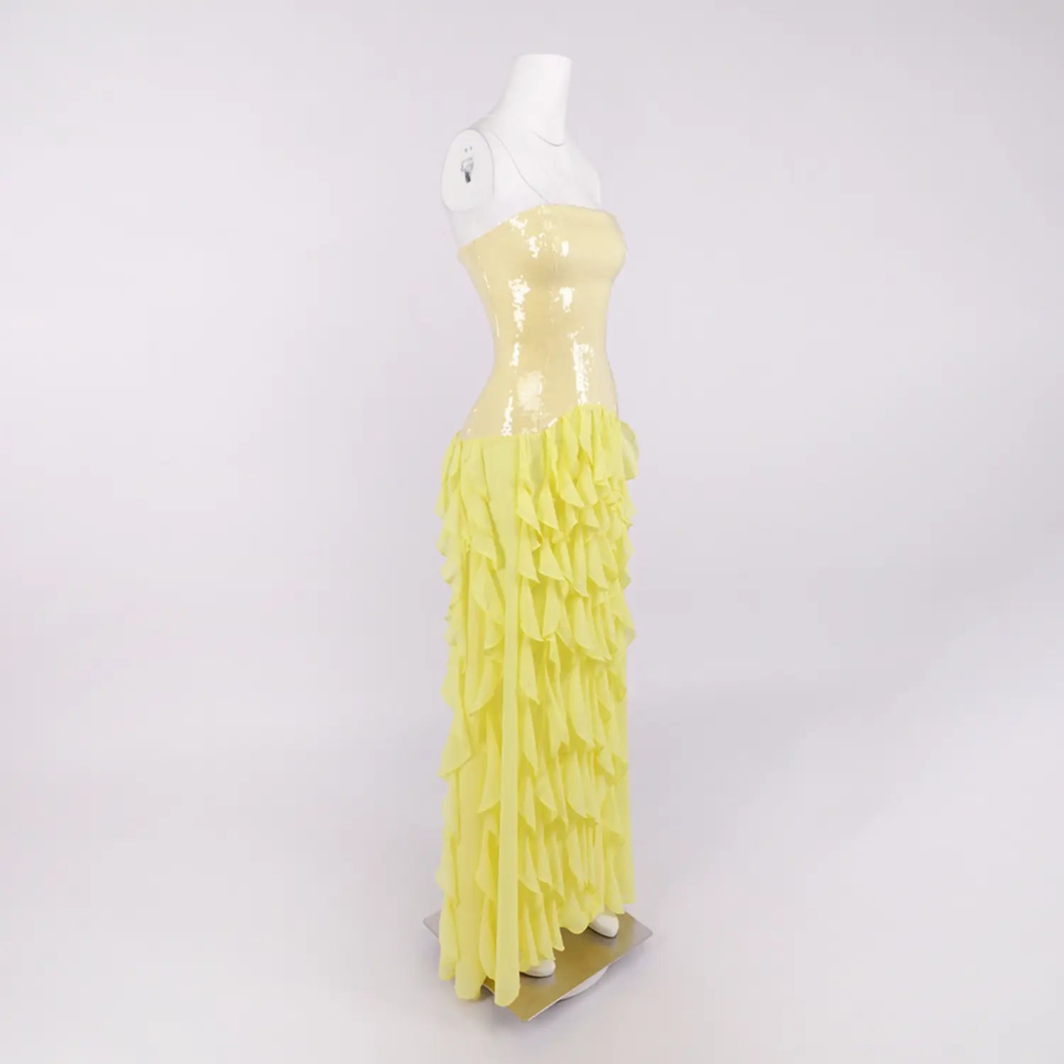 Side profile white studio shot of yellow strapless maxi gown, slim sequin torso, asymmetrical layered chiffon ruffles and high side skirt slit