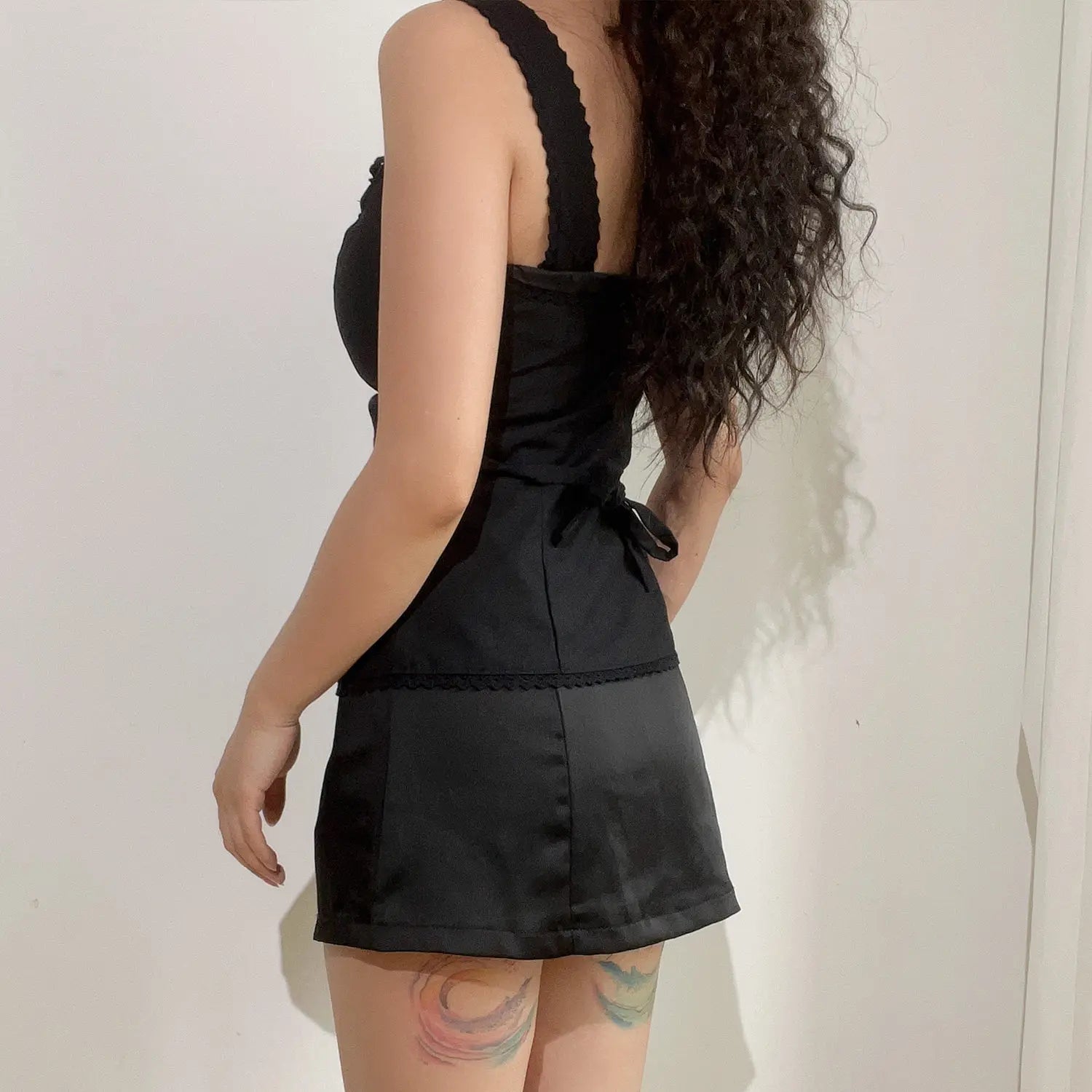 Side back lifestyle view of black two piece set, scallop edged cami corset top with rear waist tie sash, paired with smooth glossy satin mini skirt