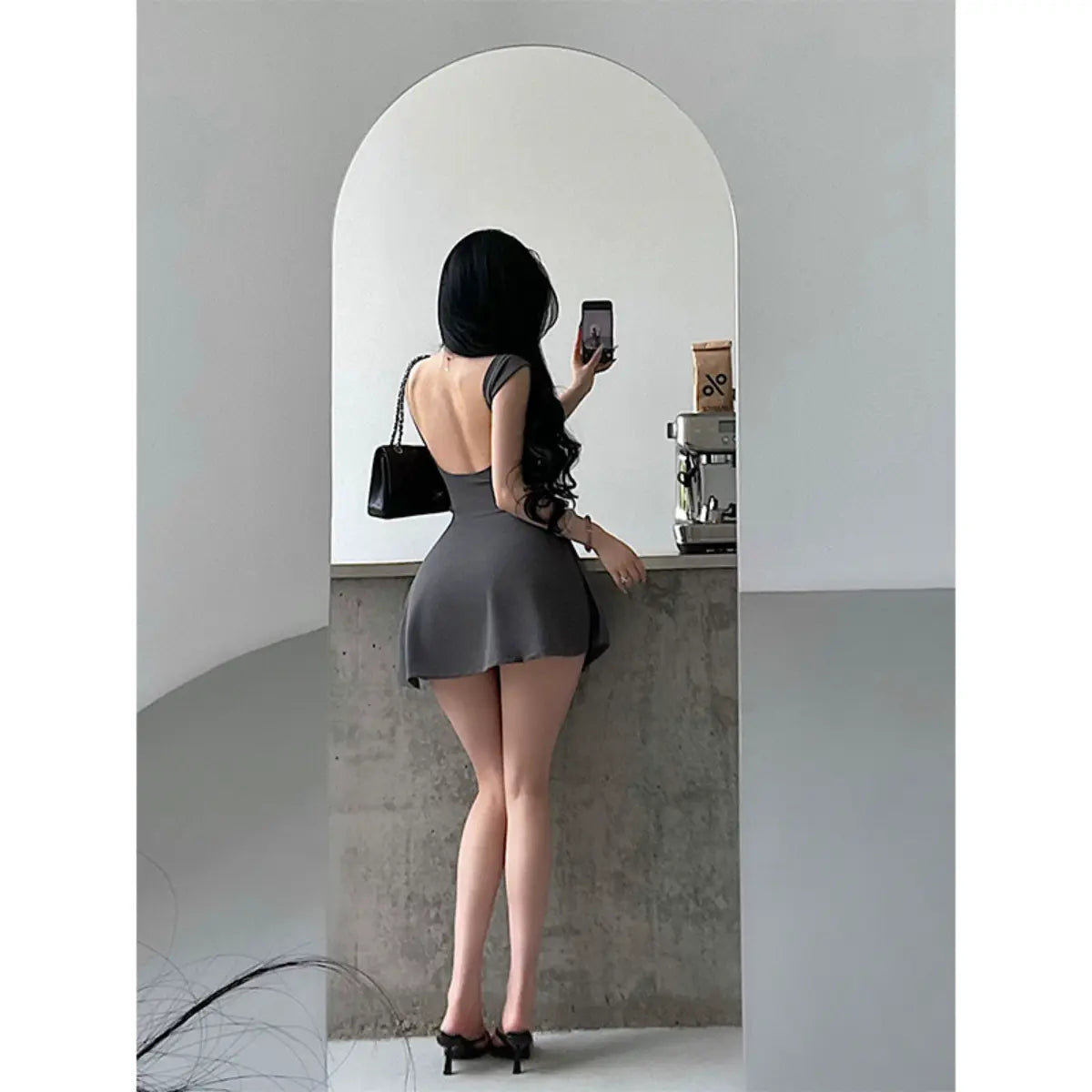 Back view of a woman wearing a gray square-neck, low-back fit-and-flare mini dress taking a mirror selfie.
