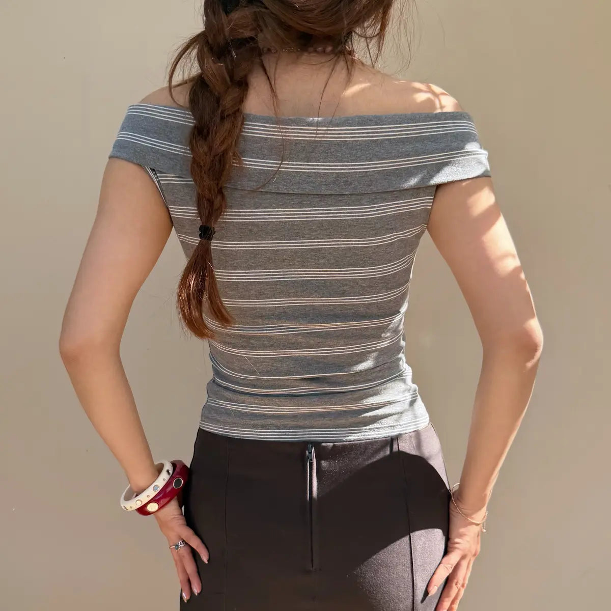  Back view of a woman wearing a gray striped off-shoulder top with a folded neckline, paired with a black skirt.