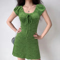 Indoor lifestyle full shot of model wearing bright green embossed ginkgo mini dress, front bust tie detail, styled with delicate blue pendant necklace and small silver shoulder bag