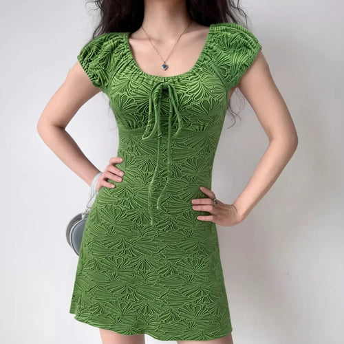 Indoor lifestyle full shot of model wearing bright green embossed ginkgo mini dress, front bust tie detail, styled with delicate blue pendant necklace and small silver shoulder bag