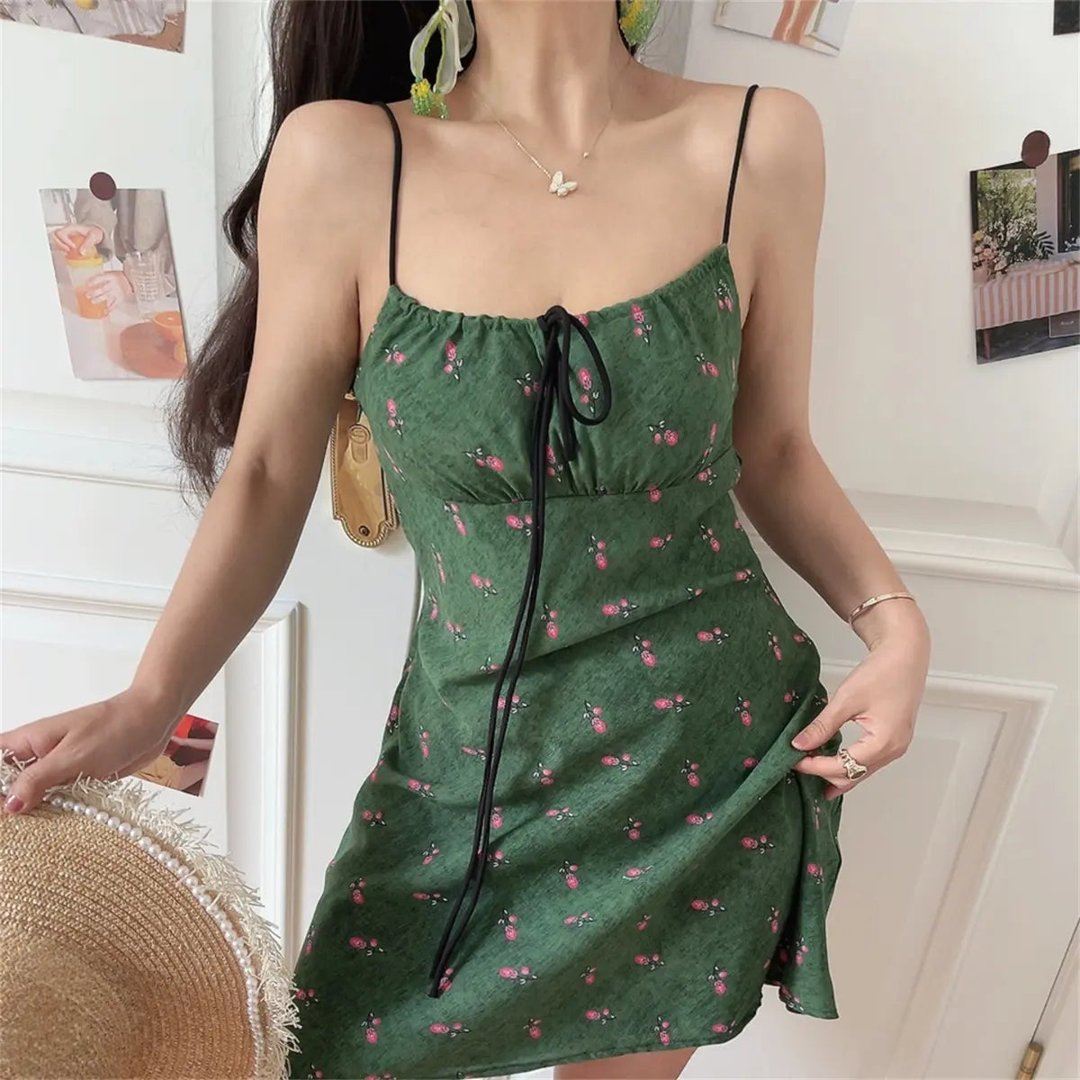 Close-up view of the front of a woman wearing a green floral-print spaghetti-strap dress with a black drawstring tie, holding a straw hat.