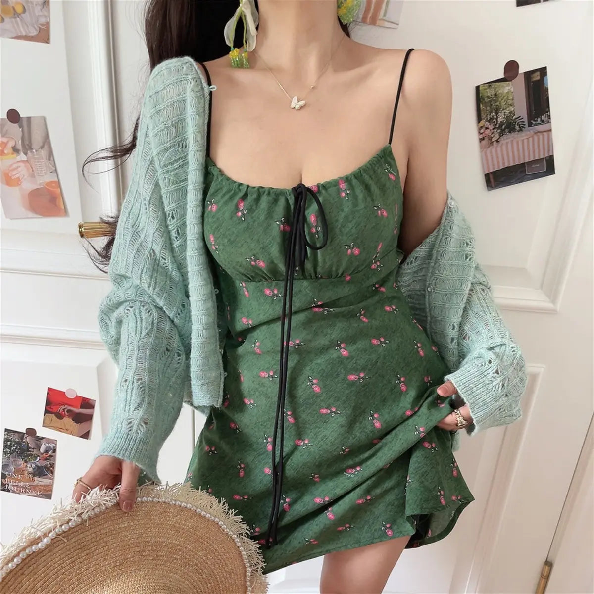 Woman wearing a green floral-print spaghetti-strap dress with a black drawstring tie, paired with a light blue knit cardigan, holding a straw hat.