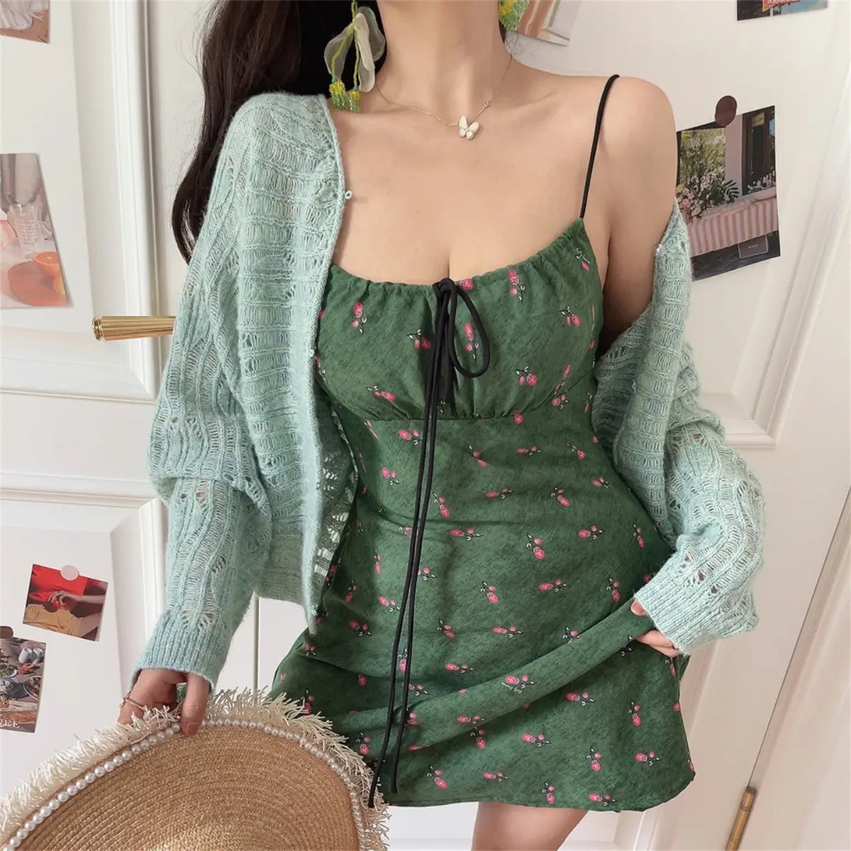 Woman wearing a green floral-print spaghetti-strap dress with a black drawstring tie, paired with a light blue knit cardigan, holding a straw hat.