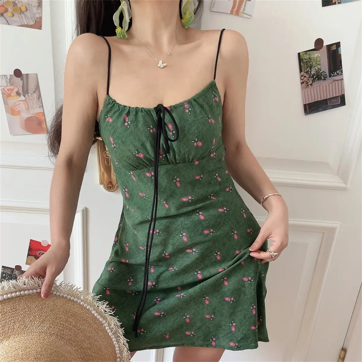 Woman wearing a green floral-print spaghetti-strap dress with a black drawstring tie, holding a straw hat and a gold bag.