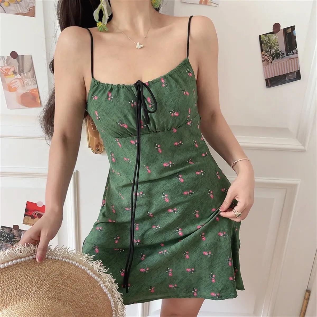 Woman wearing a green floral-print spaghetti-strap dress with a black drawstring tie, holding a straw hat and a gold bag.