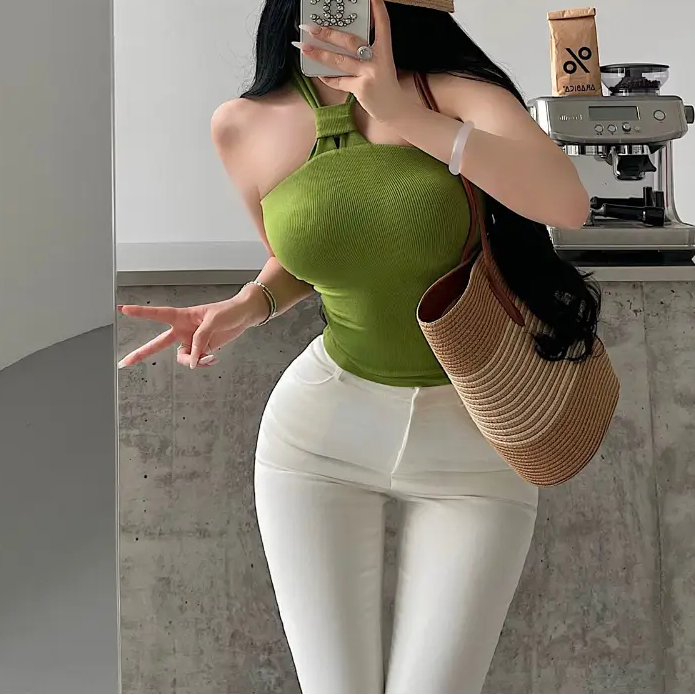 A woman wearing a green ribbed keyhole halter top, styled with white pants, a straw hat, and a woven bag, taking a mirror selfie.