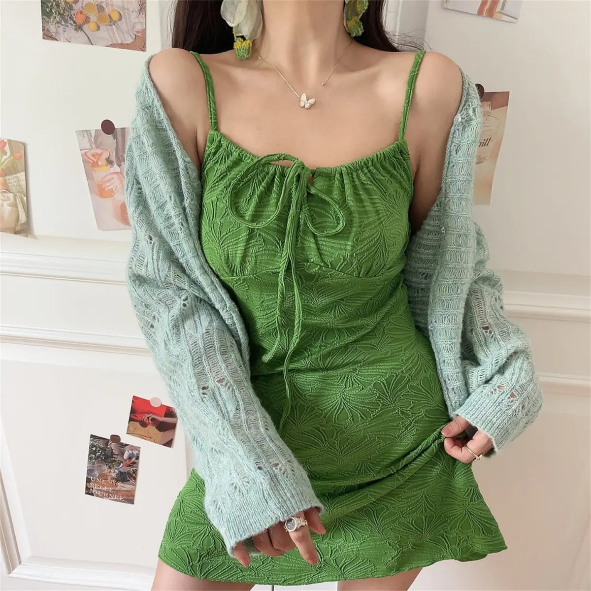 Woman wearing a green textured mini dress with spaghetti straps and front tie, layered with a light blue knit cardigan.