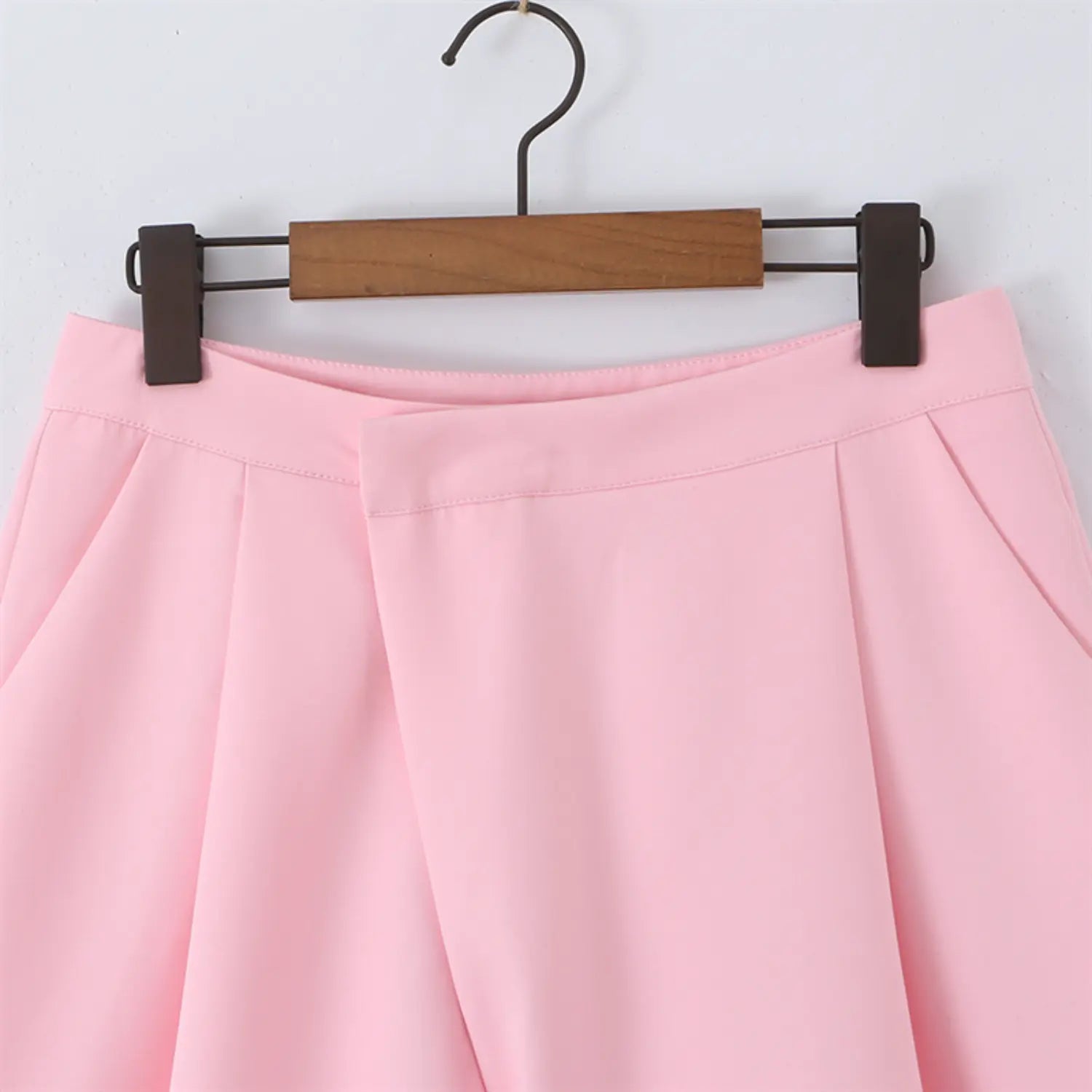 Hanger close-up of front waist, overlapping wrap panel, high waistband stitching, front fold detail, pale pink tailored fabric