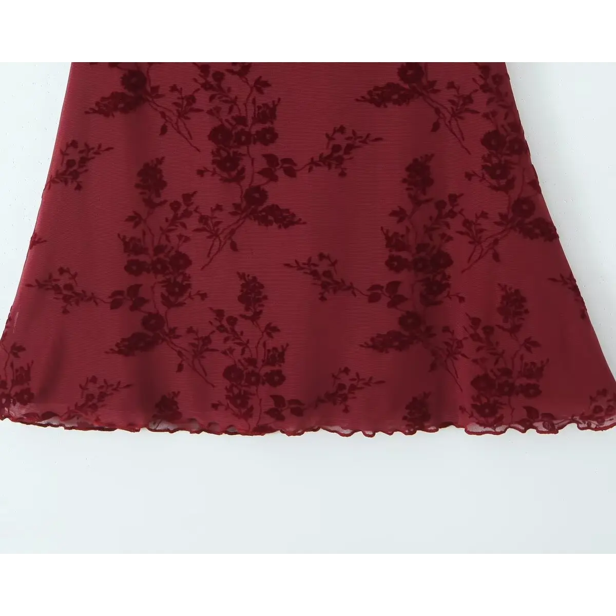 Close-up of the hem on a burgundy flocked floral mesh dress, highlighting the ruffled, scalloped edge and flocked floral pattern.