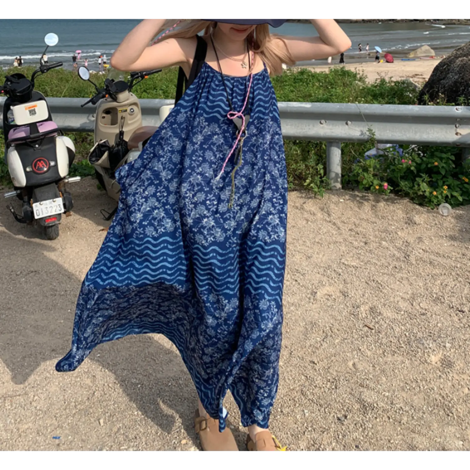 Wind-swept outdoor seaside roadside lifestyle shot of oversized batik print cami maxi dress, deep blue floral & wavy stripe pattern, thin adjustable spaghetti straps, lightweight flowy fabric billowing in wind, black shoulder tote bag, beige clogs, beach guardrail & coastal ocean backdrop