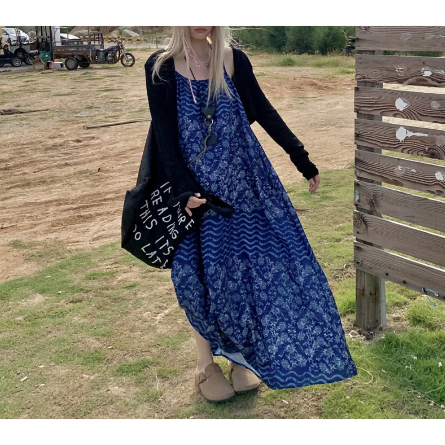 Casual outdoor grass field lifestyle shot of batik print loose maxi slip dress, layered with thin long sleeve black knit open cardigan, thin spaghetti straps, full flowy floor-grazing skirt, model carrying black letter print canvas tote bag, beige clogs, rural dirt lot & wooden fence backdrop