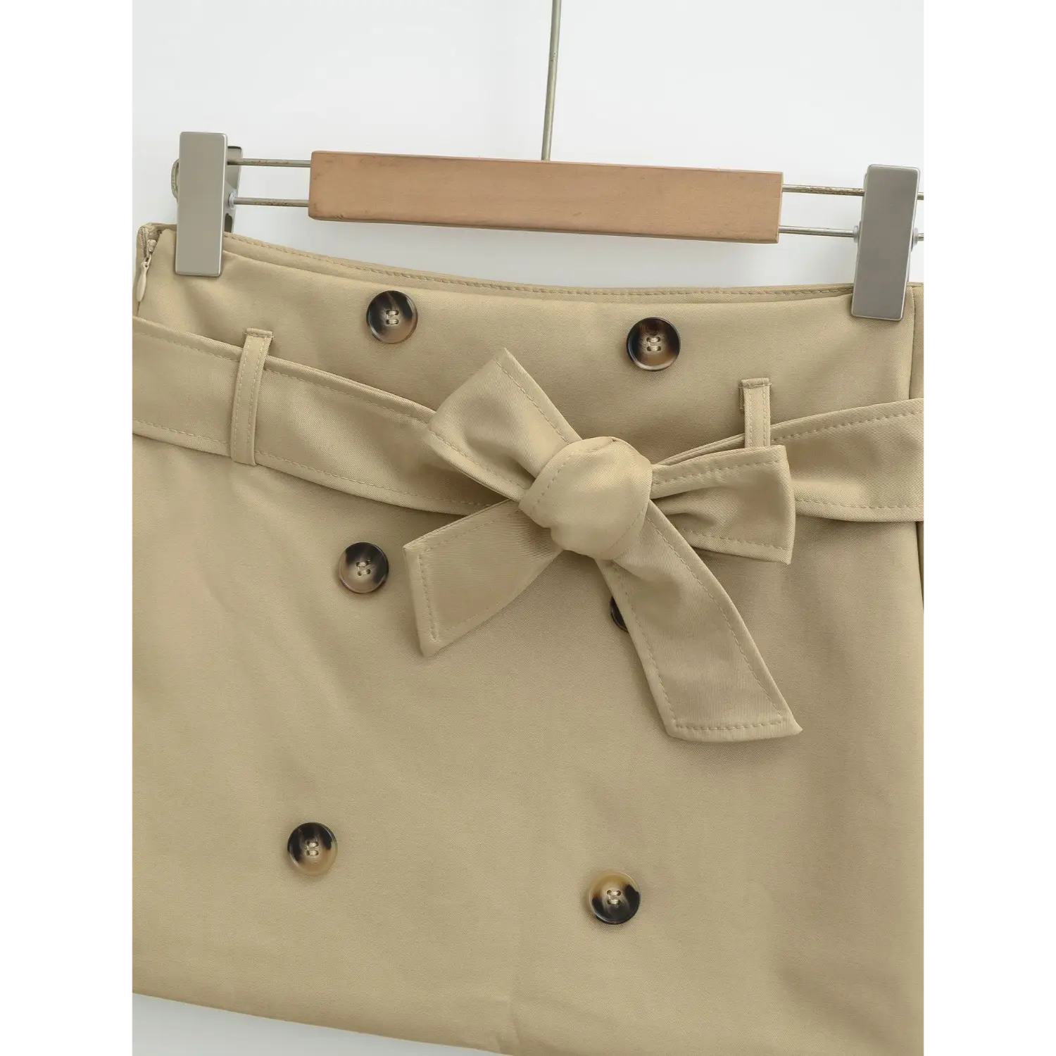Khaki mini skirt front waist macro flat close-up, three-dimensional bow tie strap symmetrical double horn buttons visible edge stitching high waist belt loops twill fabric white background detail shot