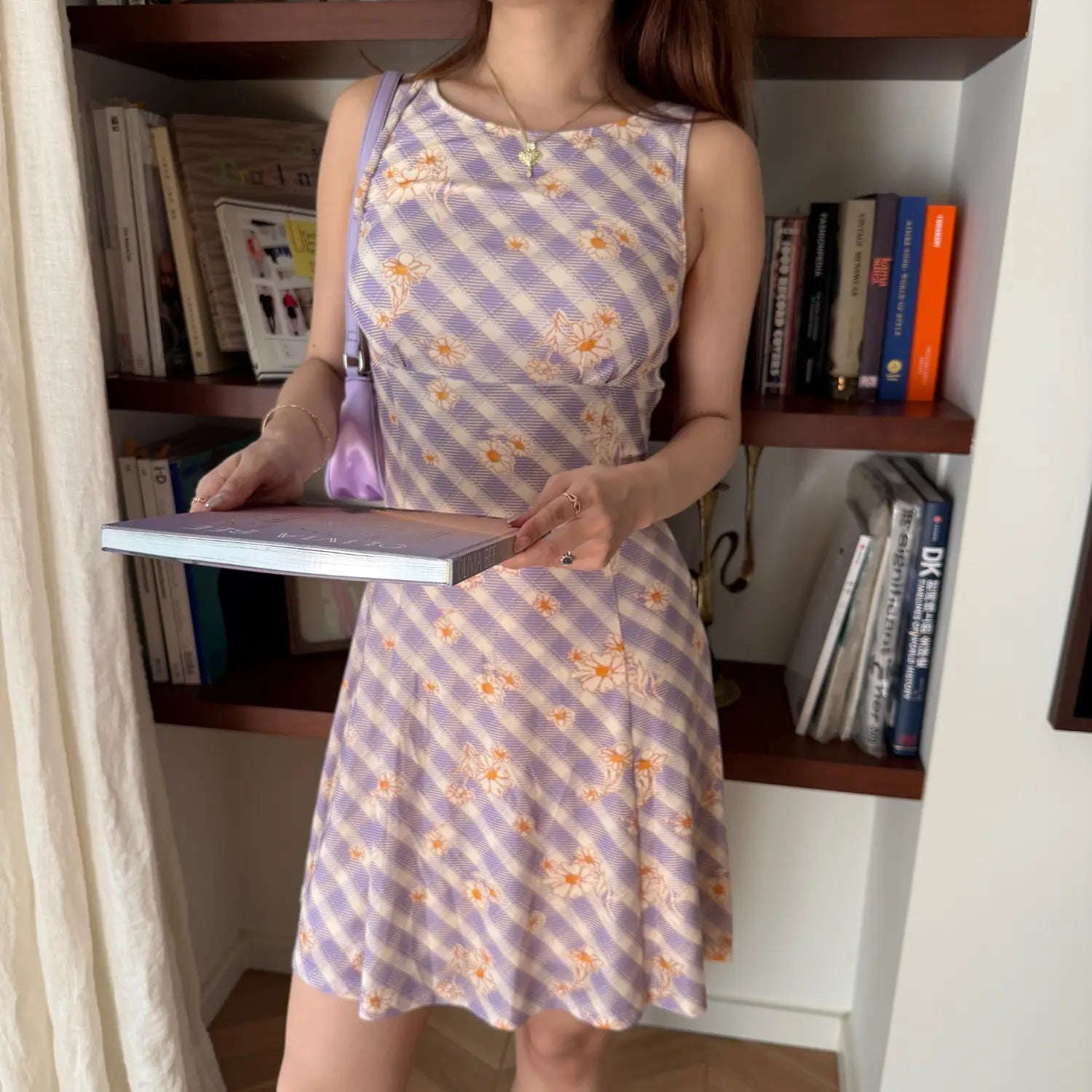  Lifestyle shot of a woman in a lavender checkered daisy print dress, holding a book in front of a bookshelf
