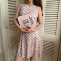 Lifestyle shot of a woman in a lavender checkered daisy print dress, holding a pink magazine in front of a louvered door
