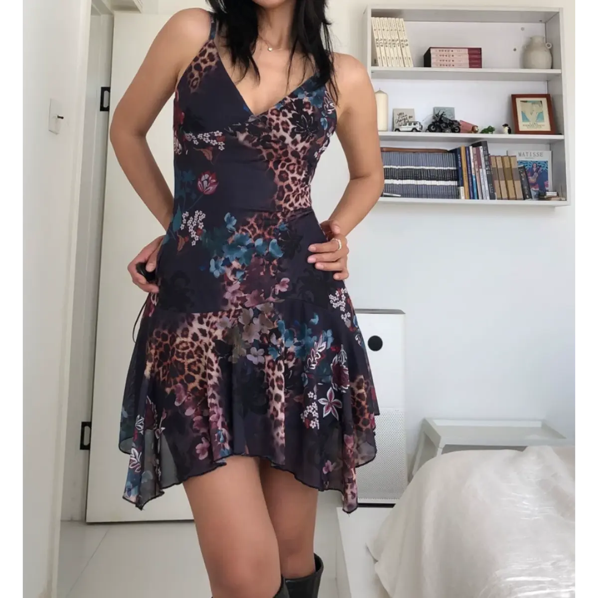 Woman wearing a form-fitting mesh mini dress with a dark leopard and floral print, spaghetti straps, V-neckline, and asymmetrical ruffled hem, posed with both hands on her hips.