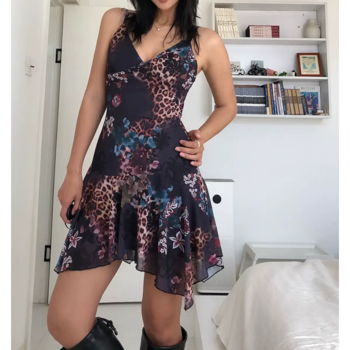 Woman wearing a form-fitting mesh mini dress with a dark leopard and floral print, spaghetti straps, V-neckline, and asymmetrical ruffled hem, posed with one hand on her hip.