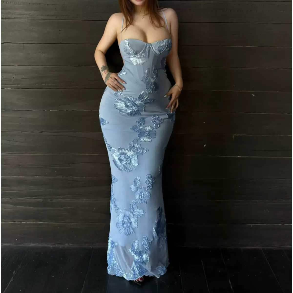 Full-body view of a woman in a light blue mermaid gown with 3D floral embroidery, posing against a dark wooden wall.