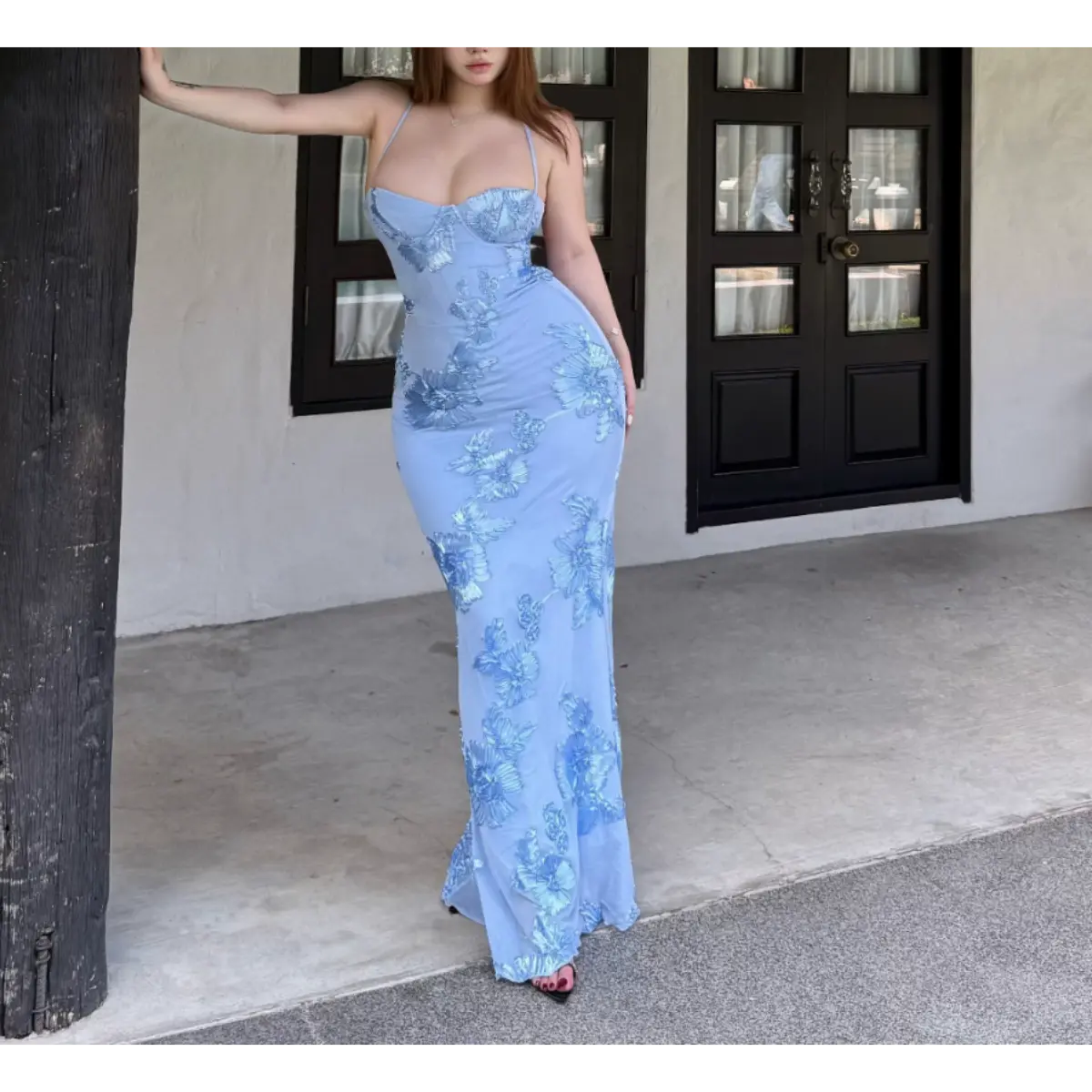 Woman in a light blue mermaid gown with 3D floral embroidery, leaning against a wooden post in front of a building entrance.