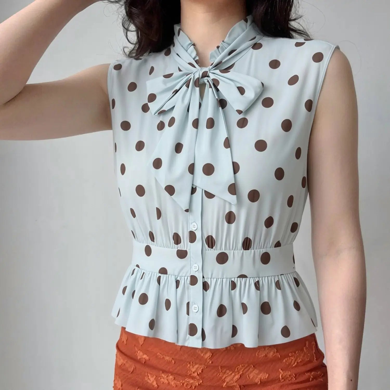 Lifestyle model outfit shot light blue polka dot sleeveless blouse, neck big bow tie, fitted smocked waist peplum hem, match with denim bottoms