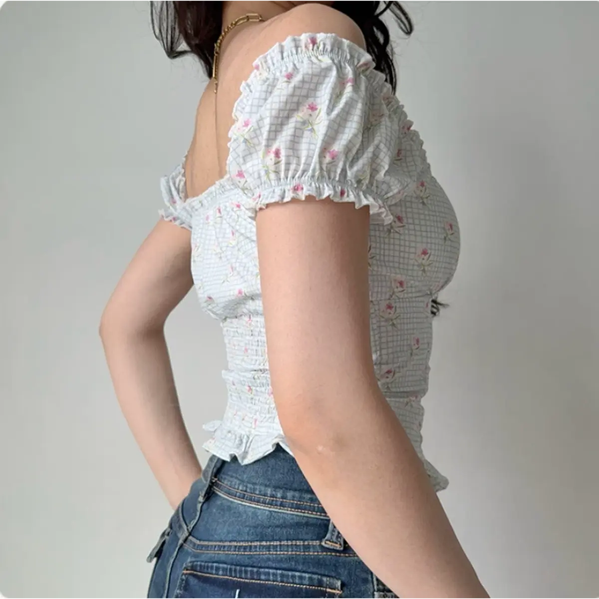 Side view of a woman wearing a light blue checkered floral puff-sleeve top with ruffled cuffs and a peplum hem, paired with jeans.