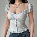 Woman in a light blue checkered floral puff-sleeve top, accessorized with a gold chain and heart pendant necklace, showing the button-front detail.