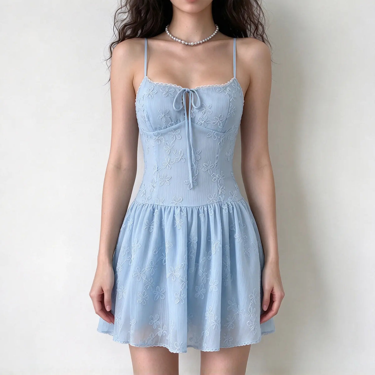 Model wearing light blue spaghetti strap mini dress, front tie detail, delicate floral embroidery, fitted waist, flowy skirt, white background