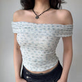Front view of a woman wearing a light blue floral off-shoulder top with a wide neckline, styled with a black flower necklace and dark jeans.