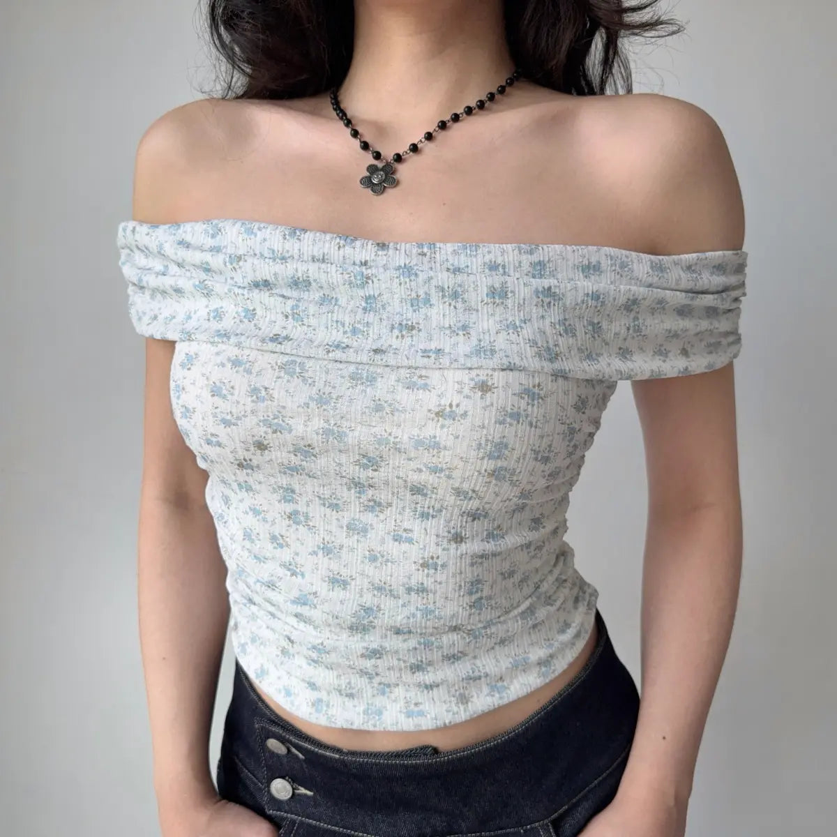 Front view of a woman wearing a light blue floral off-shoulder top with a wide neckline, styled with a black flower necklace and dark jeans.