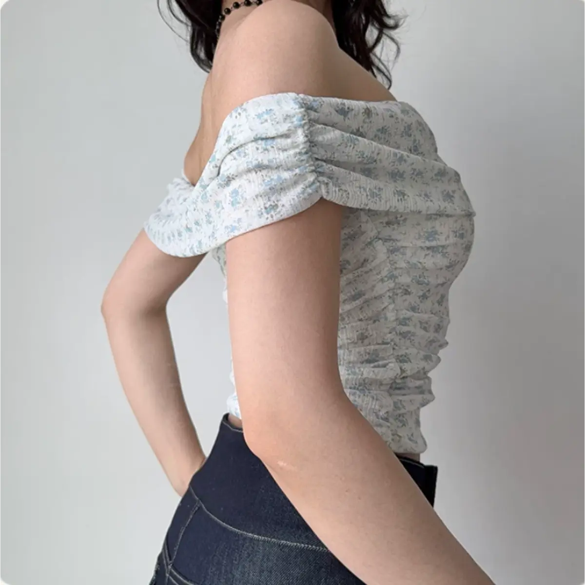  Side view of a woman wearing a light blue floral off-shoulder top with gathered sleeves and side ruching, paired with dark jeans.