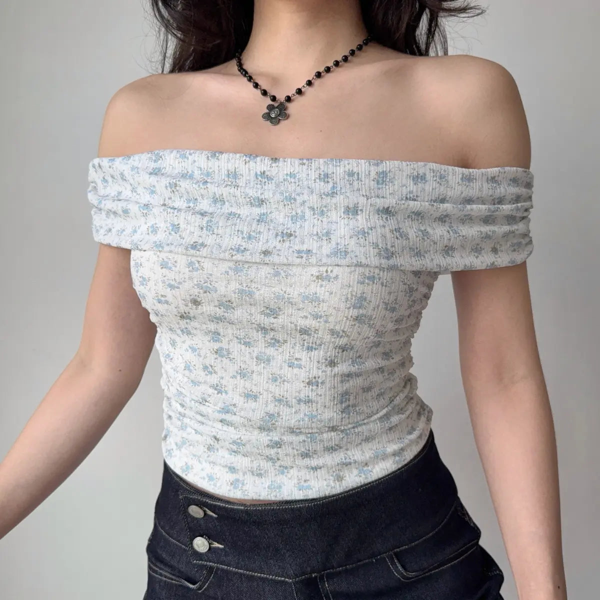 Woman wearing a light blue floral off-shoulder top, accessorized with a black flower necklace, highlighting the ribbed texture and side ruching.