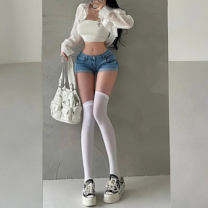Full-body shot of a woman wearing light blue low-rise denim shorts, styled with a white crop top, sheer shirt, thigh-high socks, sneakers, and a white bag.
