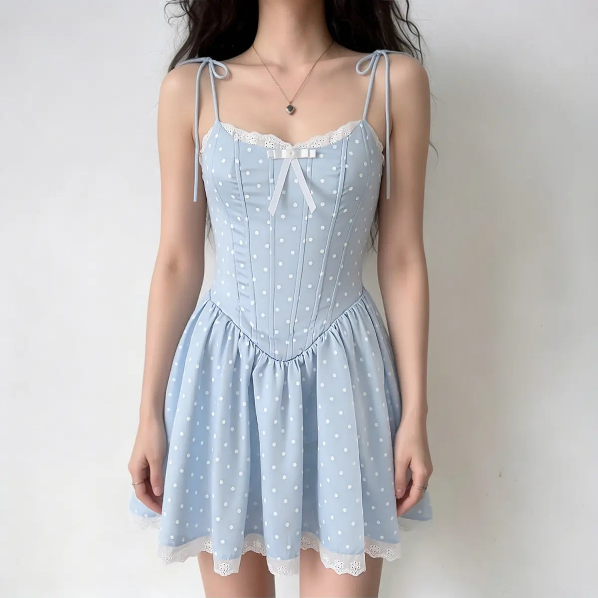 Full-body shot of a woman wearing a light blue polka dot corset-style mini dress with lace trim, a white bow, and tie straps.