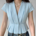 Lifestyle model wearing light blue glossy satin cropped blouse, wide lapel deep V neck, vertical front pleats, cap short sleeves, tailored flared peplum waist hem, matched blue jeans and pearl star pendant necklace daily outfit display