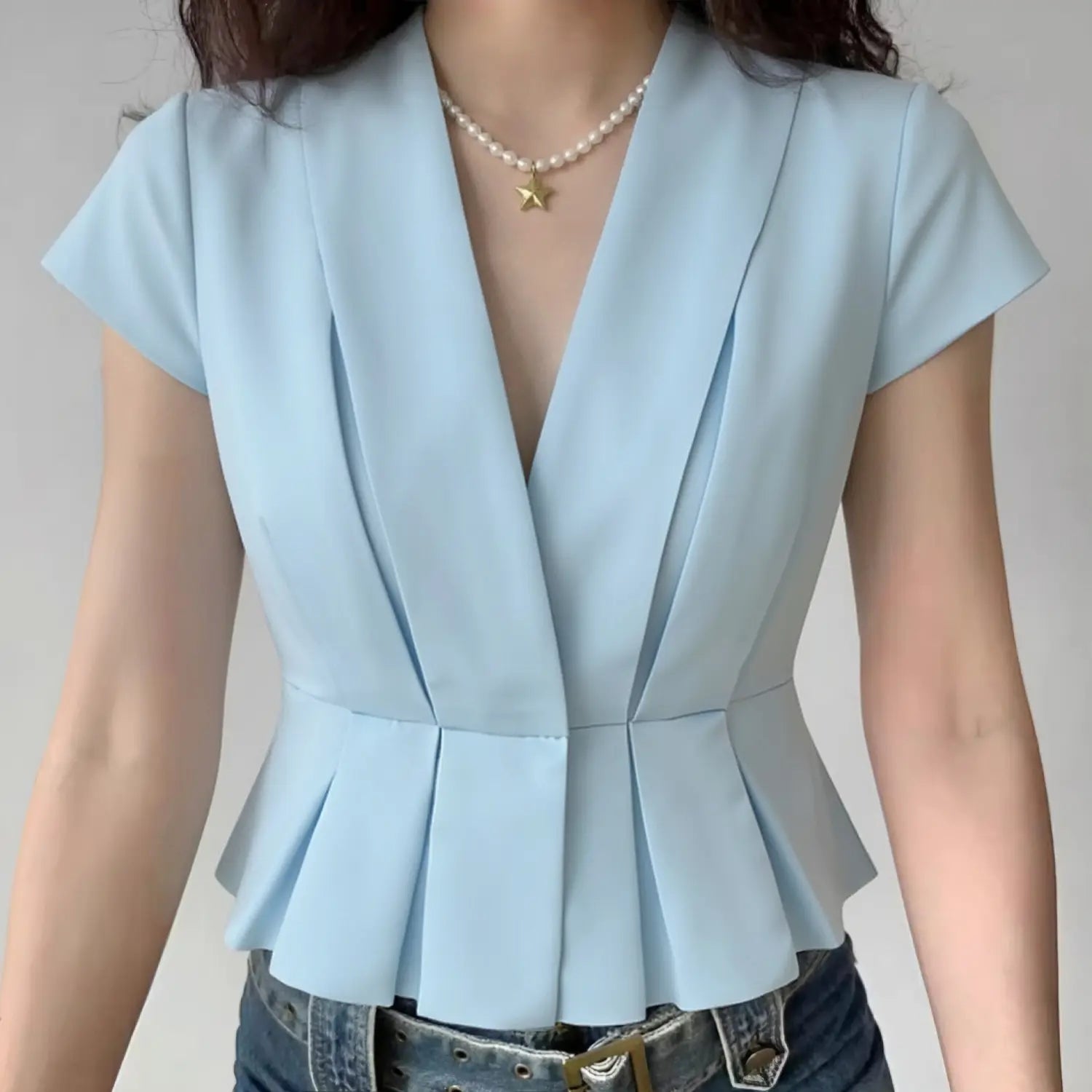 Lifestyle model wearing light blue glossy satin cropped blouse, wide lapel deep V neck, vertical front pleats, cap short sleeves, tailored flared peplum waist hem, matched blue jeans and pearl star pendant necklace daily outfit display