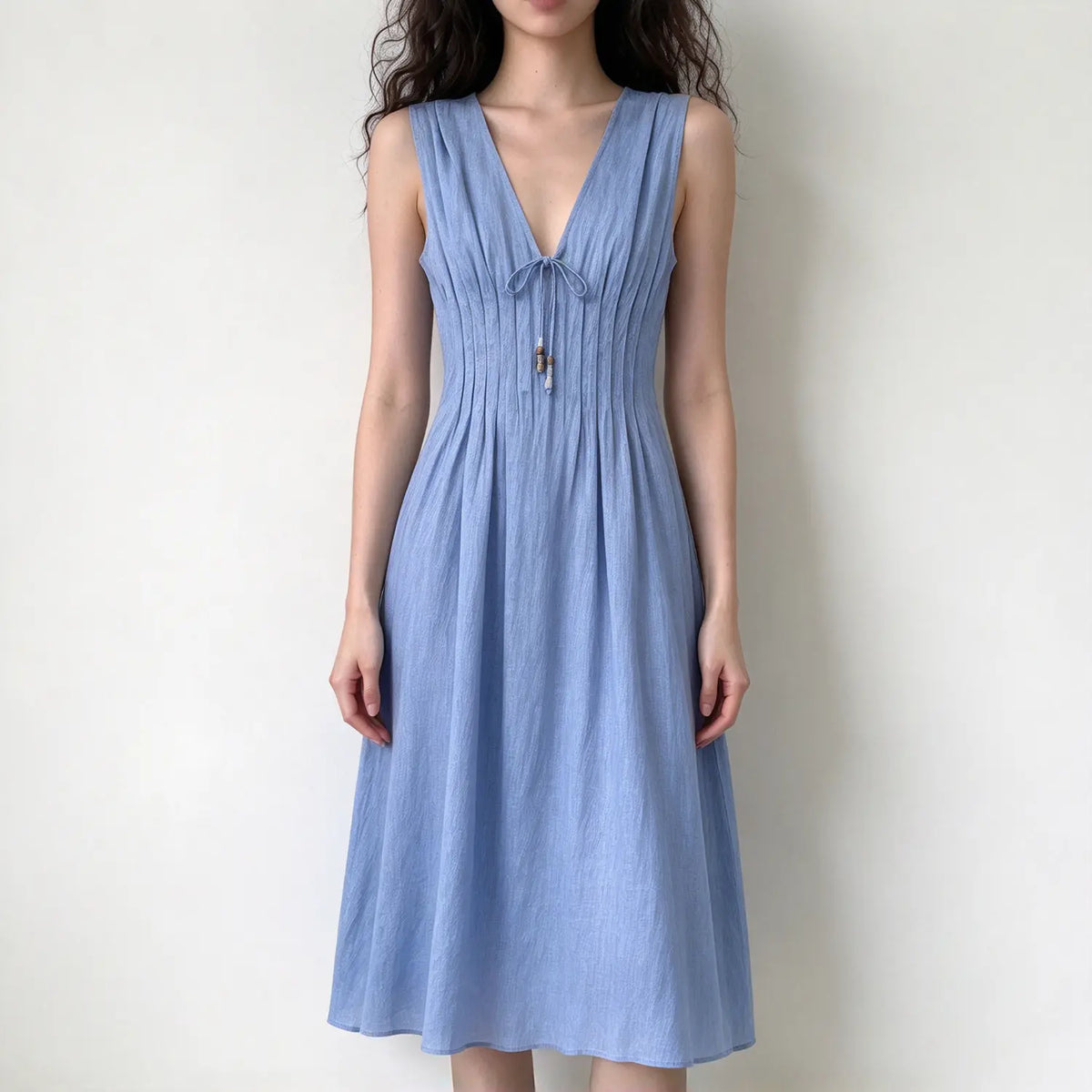 Model wearing light blue sleeveless midi dress, deep v neck with tie and bead tassel, front pleated bodice, flowy A-line skirt, white backdrop