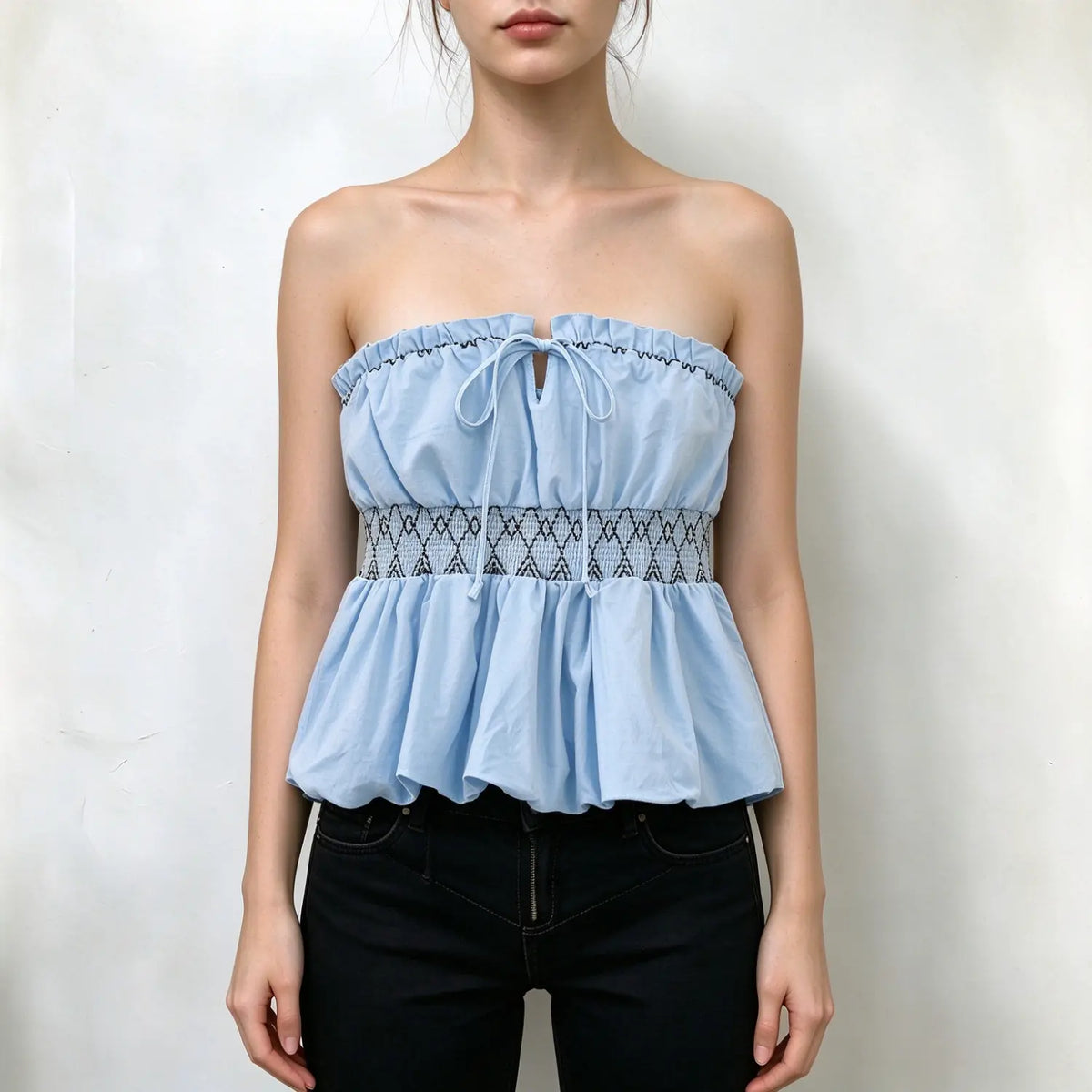 Model wearing light blue strapless tube top, ruffled neckline with front tie keyhole, geometric diamond smocked waistband, flared peplum hem, paired with black jeans